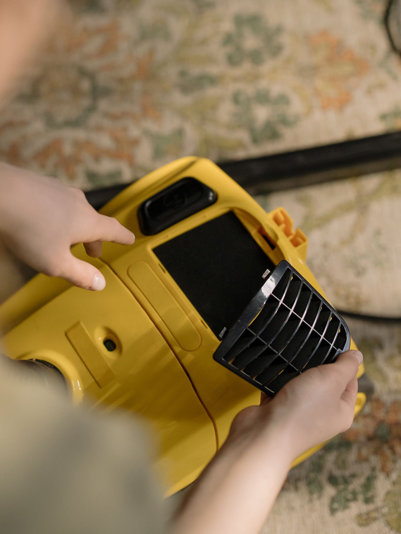 Hands removing a black filter cover from a yellow vacuum cleaner.